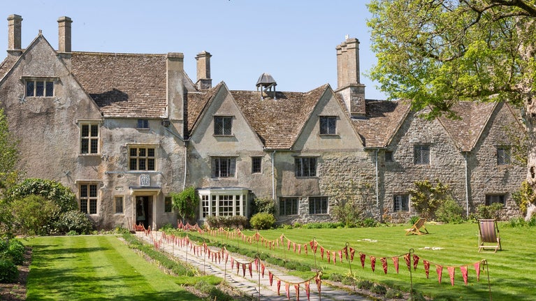 A wide photo of Avebury Manor taken from the end of the Lavender Walk on the East Lawn, flanked by green trees and a bright blue sky.